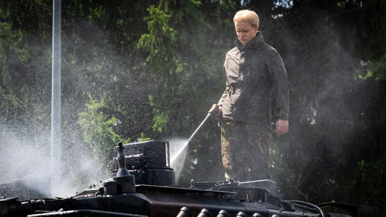 Een soldaat in een donkere jas spuit met een hogedrukreiniger water op een pantservoertuig in een bosrijke omgeving.