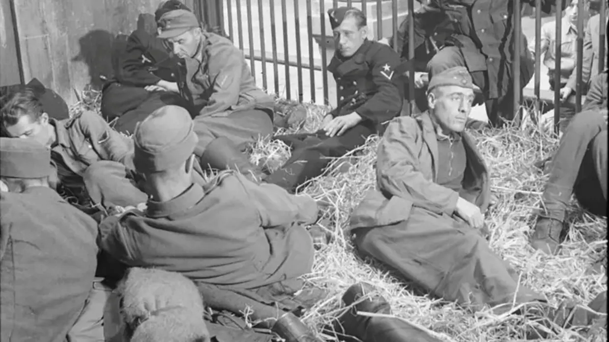 Zes mannen in militaire kleding zitten op stro, leunend tegen een houten hek in een zwart-wit foto.