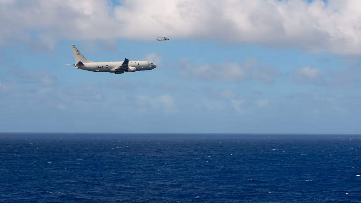 Een groot militair vliegtuig vliegt met de zee onder en blauwe lucht boven. 