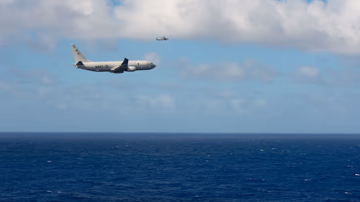 Een groot militair vliegtuig vliegt met de zee onder en blauwe lucht boven. 