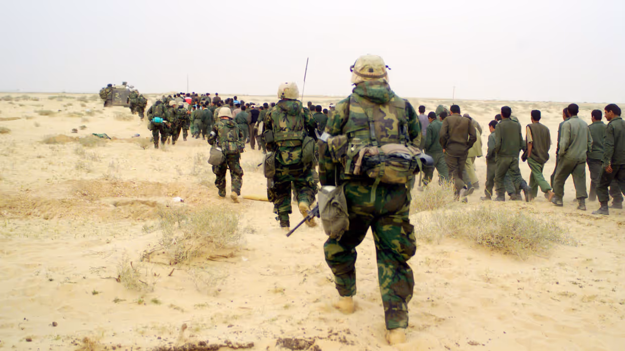 +alttekst: Een groep soldaten en burgers loopt door een zanderig, dor woestijnlandschap, met militaire voertuigen en uitrusting op de achtergrond. 