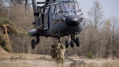 Een militaire helikopter landt in een stoffige, open vlakte. Twee soldaten staan op de grond en wijzen naar de helikopter. 