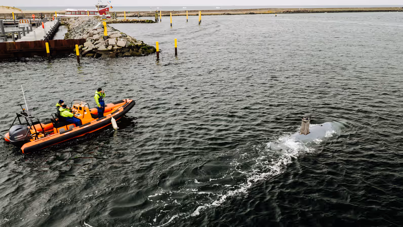Een oranje reddingsboot met drie opvarenden vaart op zee, terwijl een kleine onderzeeër naast hen door het water beweegt.
