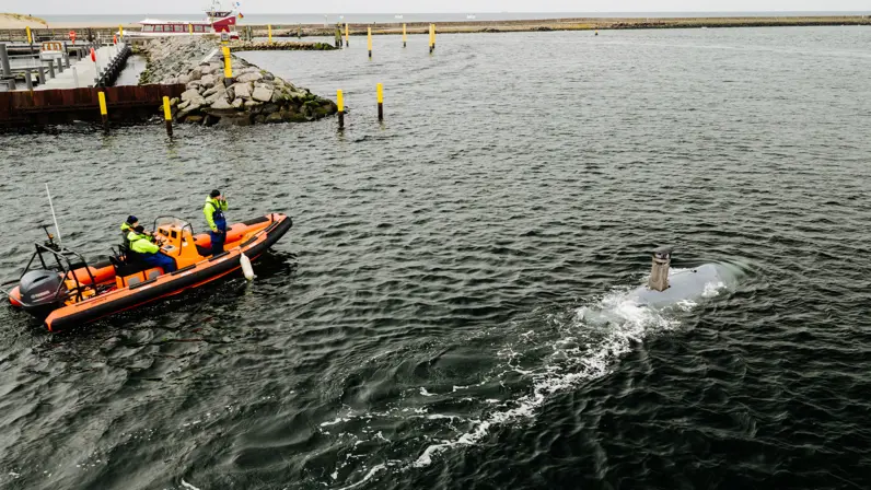 Een oranje reddingsboot met drie opvarenden vaart op zee, terwijl een kleine onderzeeër naast hen door het water beweegt.