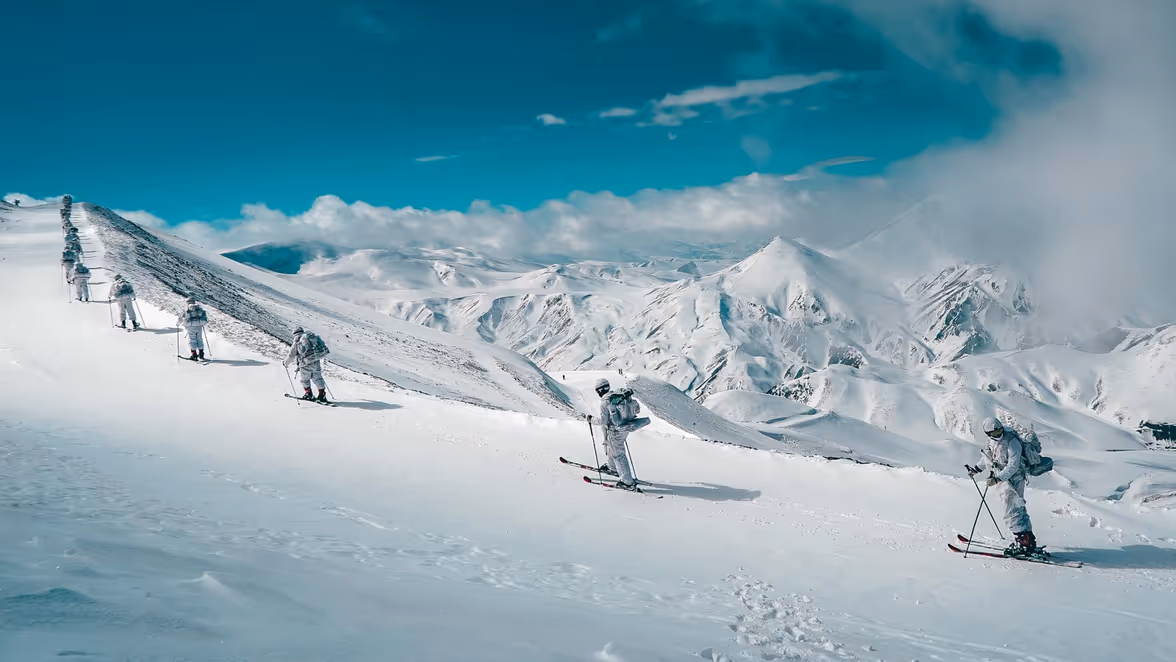 Vijf mensen in witte winterkleding skiën in een rij over een besneeuwde berghelling met indrukwekkende, besneeuwde bergen en een blauwe lucht op de achtergrond. 