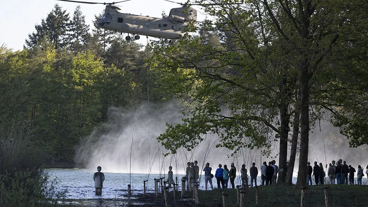 Helikopter schept water op uit een meer, groene bomen, toeschouwers kijken toe vanaf de oever in avondlicht. 