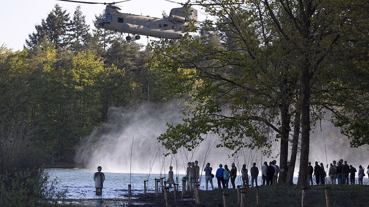 Helikopter schept water op uit een meer, groene bomen, toeschouwers kijken toe vanaf de oever in avondlicht. 