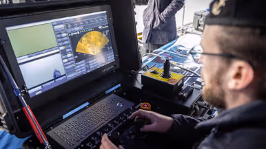 Een marinecollega zit gebogen op de kade en is bezig met een geel vaartuigje dat half onder water ligt. Op de andere foto bedient een marinecollega joysticks en kijkt naar een beeldscherm waarop onderwaterbeelden zijn te zien. 