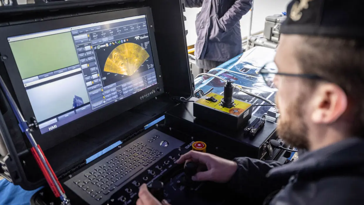 Een marinecollega zit gebogen op de kade en is bezig met een geel vaartuigje dat half onder water ligt. Op de andere foto bedient een marinecollega joysticks en kijkt naar een beeldscherm waarop onderwaterbeelden zijn te zien. 
