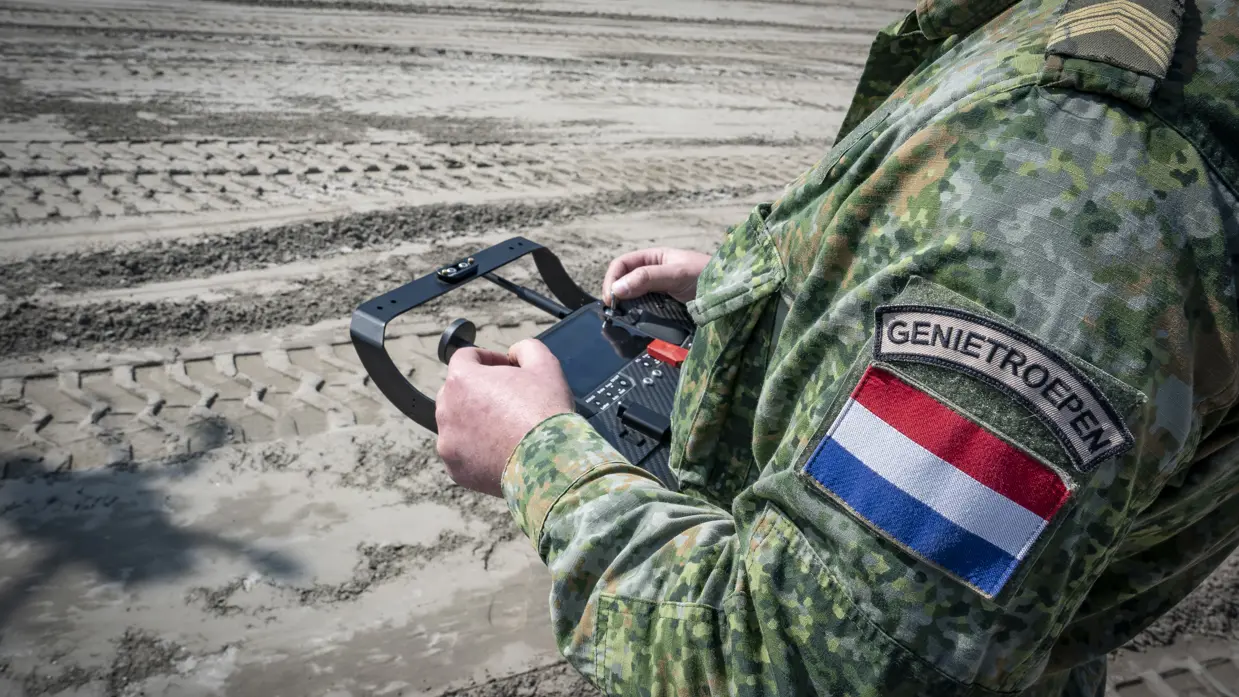  Een militair in groen pak bestuurt een drone. Op de mouw staat de tekst ‘Genietroepen’.
