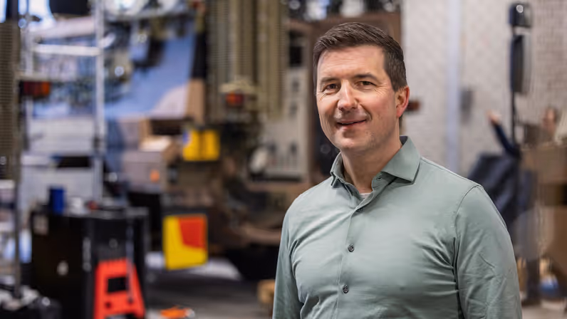 Een gecamoufleerde militair houdt op een knie gewapend de wacht aan de achterzijde van een Bushmaster, die in een groene strook van bos en struik geparkeerd staat. Op de Bushmaster zijn diverse antennes te zien. Foto rechts: Niels kijkt recht in de camera. Hij draagt een lichtgroene blouse en heeft kort donker haar.