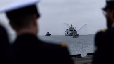 Marineschip de ‘Vlissingen’ op zee, daarnaast twee sleepboten die met hun waterkanonnen spuiten. Het schip wordt voorgegaan door een kleine mijnenveger. Op de tweede afbeelding hetzelfde schip, maar dan gezien langs een tweetal marineofficieren aan de wal. 