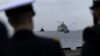 De Vlissingen met daarnaast een sleepboot, gezien vanaf de kade. Daarnaast het grijze marineschip van dichterbij, de sleepboot er nog altijd naast varend.