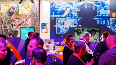 Groep mensen staat rond stand op conferentievloer. Een man wijst op computerscherm in een stand op de conferentie en een man in blauwe trui op de rug gezien zit tussen 2 mannen met VR-bril op.