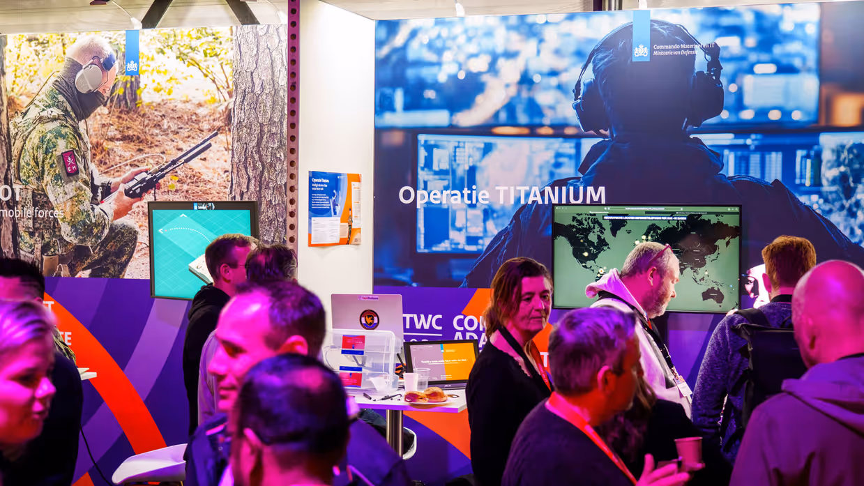 Groep mensen staat rond stand op conferentievloer. Een man wijst op computerscherm in een stand op de conferentie en een man in blauwe trui op de rug gezien zit tussen 2 mannen met VR-bril op.