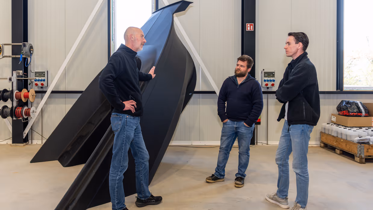 Drie mensen in fabriekshal in gesprek bij een onderdeel van 3D-geprinte boot.