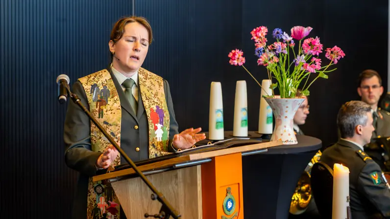 Een militair dominee spreekt vanachter een microfoon en maakt daarbij gebruik van haar handen. Ze heeft haar ogen gesloten.&nbsp;