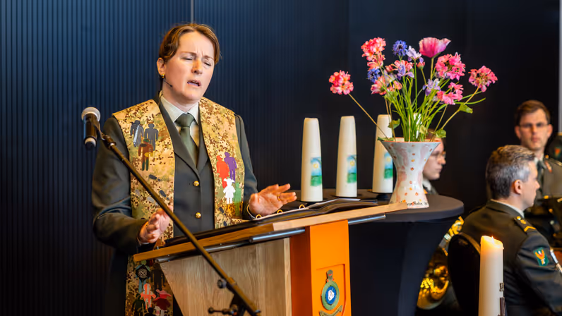 Een militair dominee spreekt vanachter een microfoon en maakt daarbij gebruik van haar handen. Ze heeft haar ogen gesloten.&nbsp;