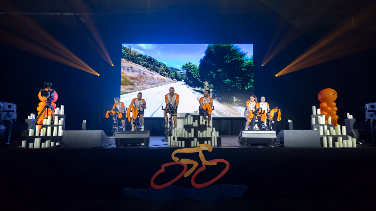 Deelnemers van team Marechaussee aan de Alpe d’HuZes Indoorride 2026 op een podium met beeldscherm erachter. 