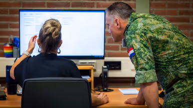 Een vrouw met opgestoken haar zit aan een bureau achter een beeldscherm, met naast haar een gebogen, staande militair.