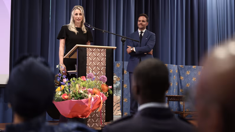 Amber, in zwarte jurk, staat achter een microfoon en spreekt de aanwezigen toe.