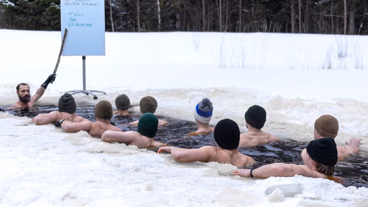 Een groep mensen zit in een ijsbad, terwijl een man met een stok instructies geeft bij een flip-over bord.