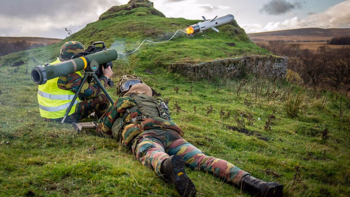 Een militair zit op de grond en vuurt vanaf zijn schouder een raket af. Deze komt met een dun touw erachter uit de afvuureenheid. Daarnaast ligt een militair op het grasveld. De twee volgen de baan van het projectiel.
