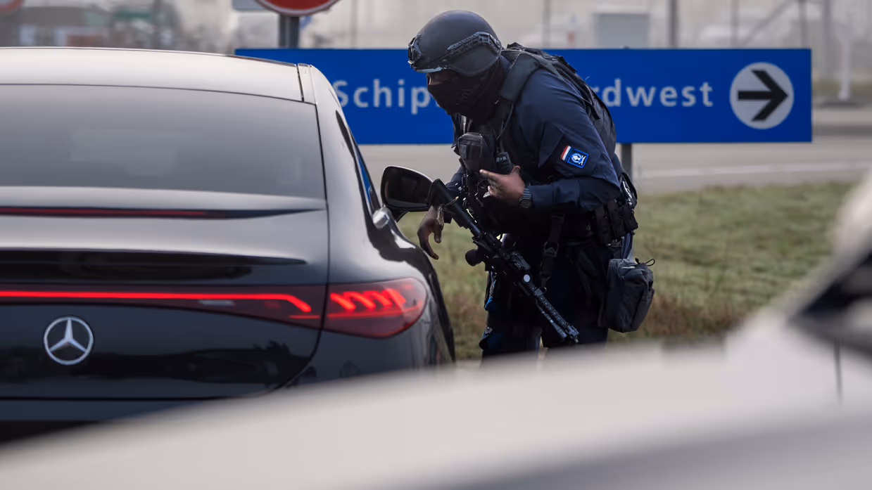 Een bewapende marechaussee in donker uniform buigt bij een zwarte Mercedes, met een blauw bord "Schiphol Noordwest" en een snelheidsbeperkingsbord op de achtergrond.