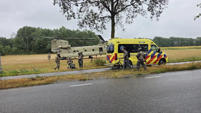 Een helikopter en een gele ambulance staan op een veld, met mensen eromheen, naast een weg en bomen.