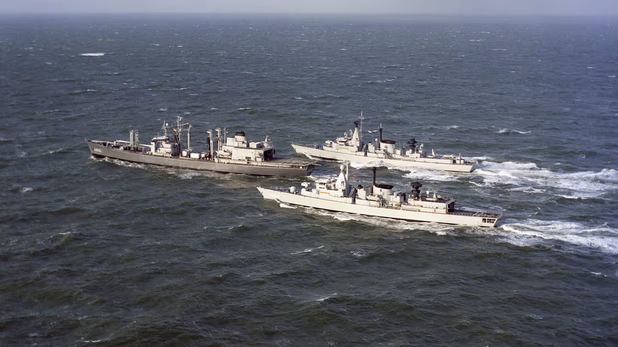Drie marineschepen varen naast elkaar over een golvende, grijze zee onder een bewolkte lucht, met antennes en apparatuur op hun dekken.