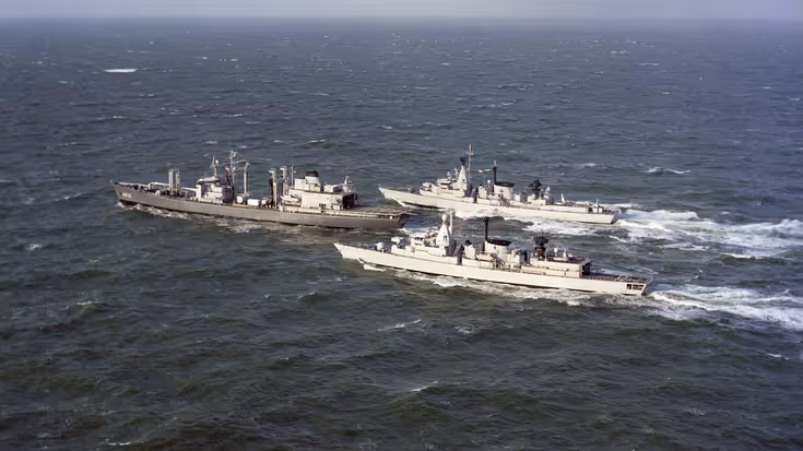 Drie marineschepen varen naast elkaar over een golvende, grijze zee onder een bewolkte lucht, met antennes en apparatuur op hun dekken.