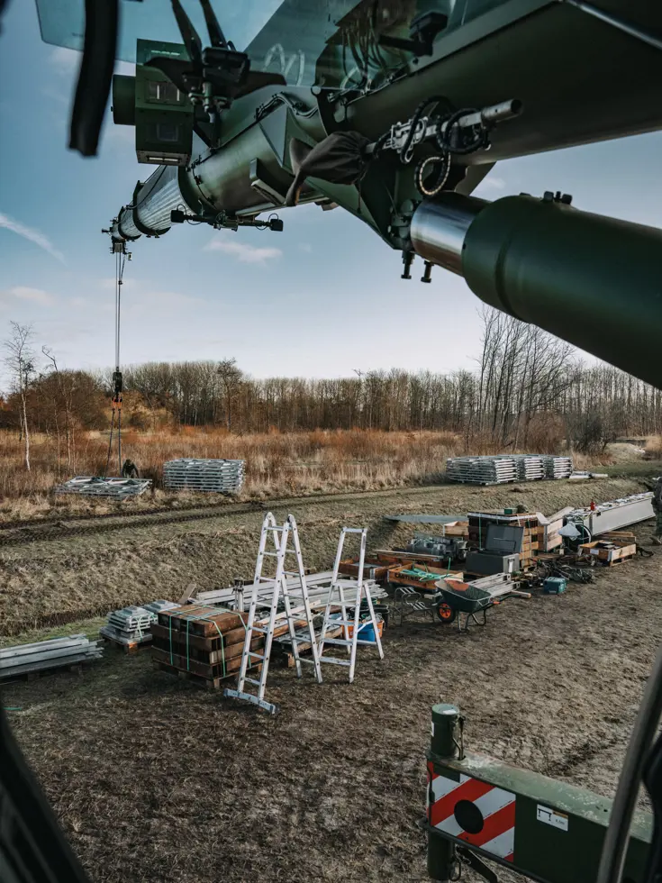 Het uitzicht vanuit een voertuig of machine met draaiende rotorbladen, gericht op een bouwplaats met materialen, ladders, kisten en stapels platen op een veld. 