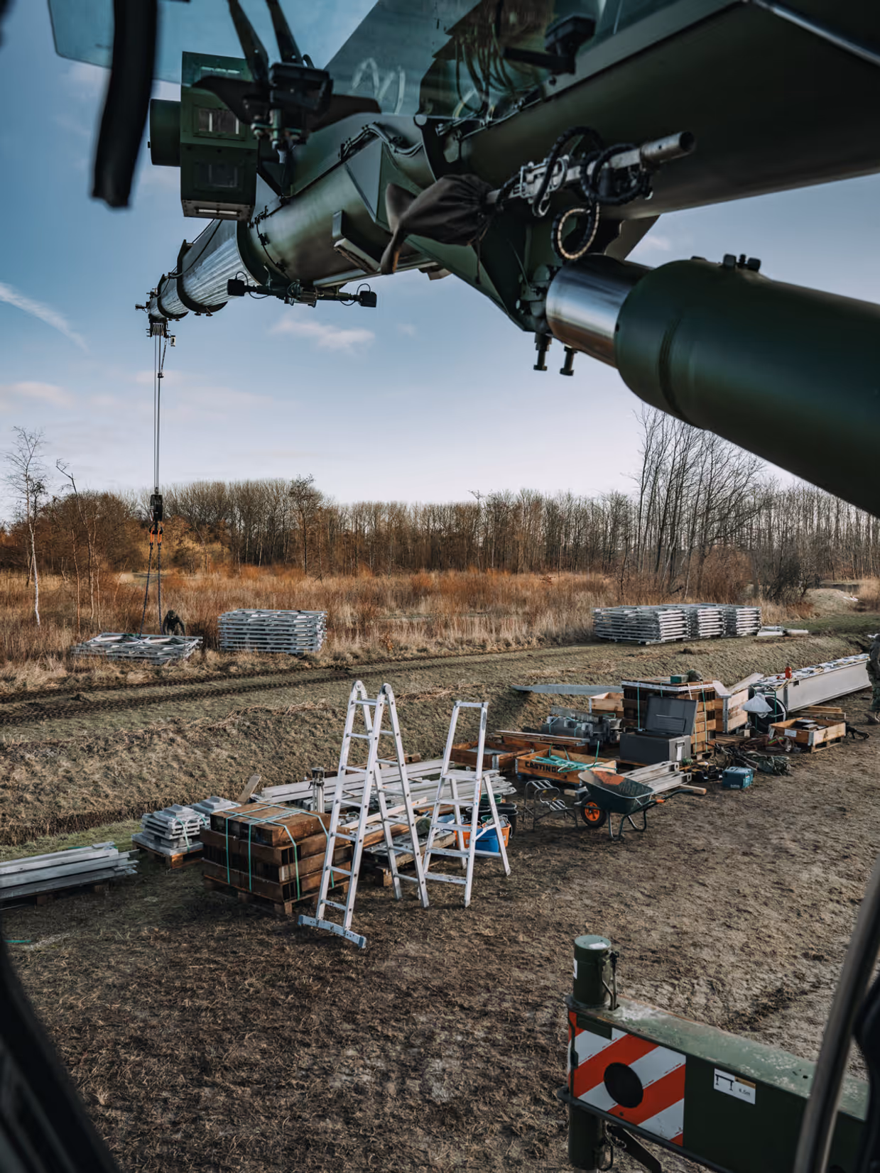 Het uitzicht vanuit een voertuig of machine met draaiende rotorbladen, gericht op een bouwplaats met materialen, ladders, kisten en stapels platen op een veld. 