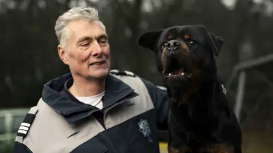 Links een man in uniform. Hij omarmt een grote zwart-bruine hond met opgetrokken oren. Achtergrond toont een bosrijke omgeving met een metalen hek. Rechts een glimlachende man in uniform staat naast een zwarte Rottweiler met open bek. Bosrijke omgeving op de achtergrond, met een ontspannen en vriendelijke sfeer. 