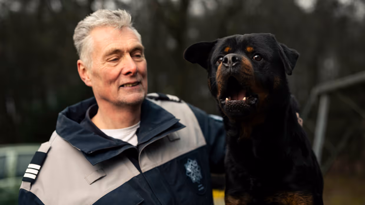 Links een man in uniform. Hij omarmt een grote zwart-bruine hond met opgetrokken oren. Achtergrond toont een bosrijke omgeving met een metalen hek. Rechts een glimlachende man in uniform staat naast een zwarte Rottweiler met open bek. Bosrijke omgeving op de achtergrond, met een ontspannen en vriendelijke sfeer. 