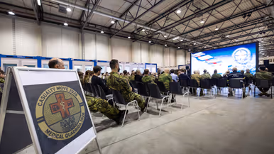  Links: Een militair in uniform staat voor een groen gecamoufleerd doek en spreekt met een microfoon. Een groep mensen in uniform en burgerkleding luistert aandachtig. Rechts: Een bord met het embleem "Casualty Move 2026 Medical Quadrigga 2026" staat op de voorgrond. Achterin zitten mensen in een grote ruimte naar een presentatie te kijken.