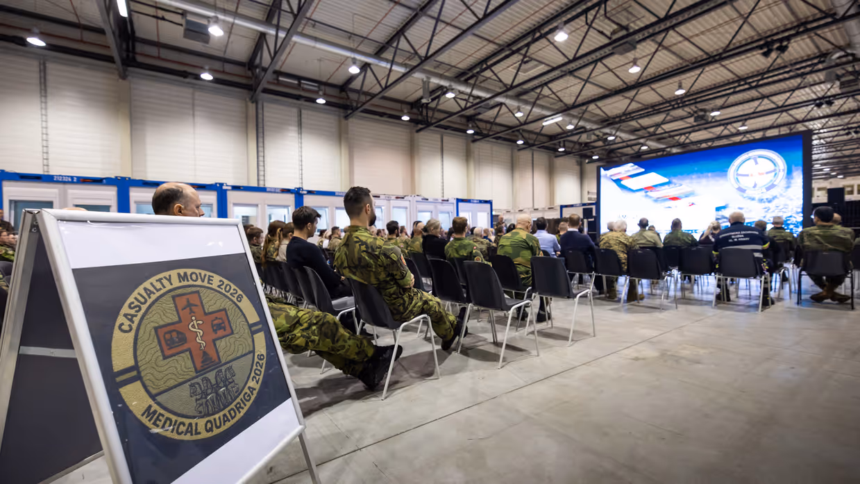  Links: Een militair in uniform staat voor een groen gecamoufleerd doek en spreekt met een microfoon. Een groep mensen in uniform en burgerkleding luistert aandachtig. Rechts: Een bord met het embleem "Casualty Move 2026 Medical Quadrigga 2026" staat op de voorgrond. Achterin zitten mensen in een grote ruimte naar een presentatie te kijken.