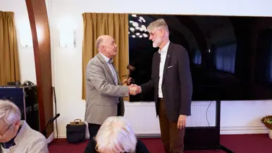 Een man in pak overhandigt een hamer aan een andere man met baard en in pak.