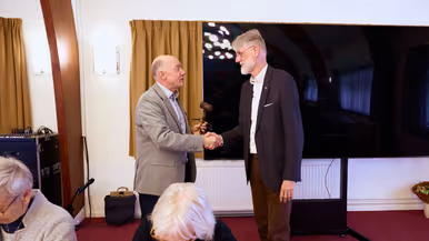 Een man in pak overhandigt een hamer aan een andere man met baard en in pak.