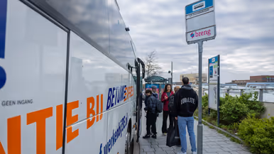 Een groep jongeren staat buiten in een rij voor een bus met "Rubens!" erop. Sommigen dragen jassen en rugzakken, een militair staat opzij. 