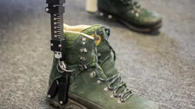 Links: een exoskelet met camouflage rugzak en groene bergschoenen staat op een tafel in een klaslokaal met stoelen en tafels.    Rechts een groene bergschoen met een zwarte metalen meetlat ernaast staat op een grijze vloer, tweede schoen op de achtergrond. 