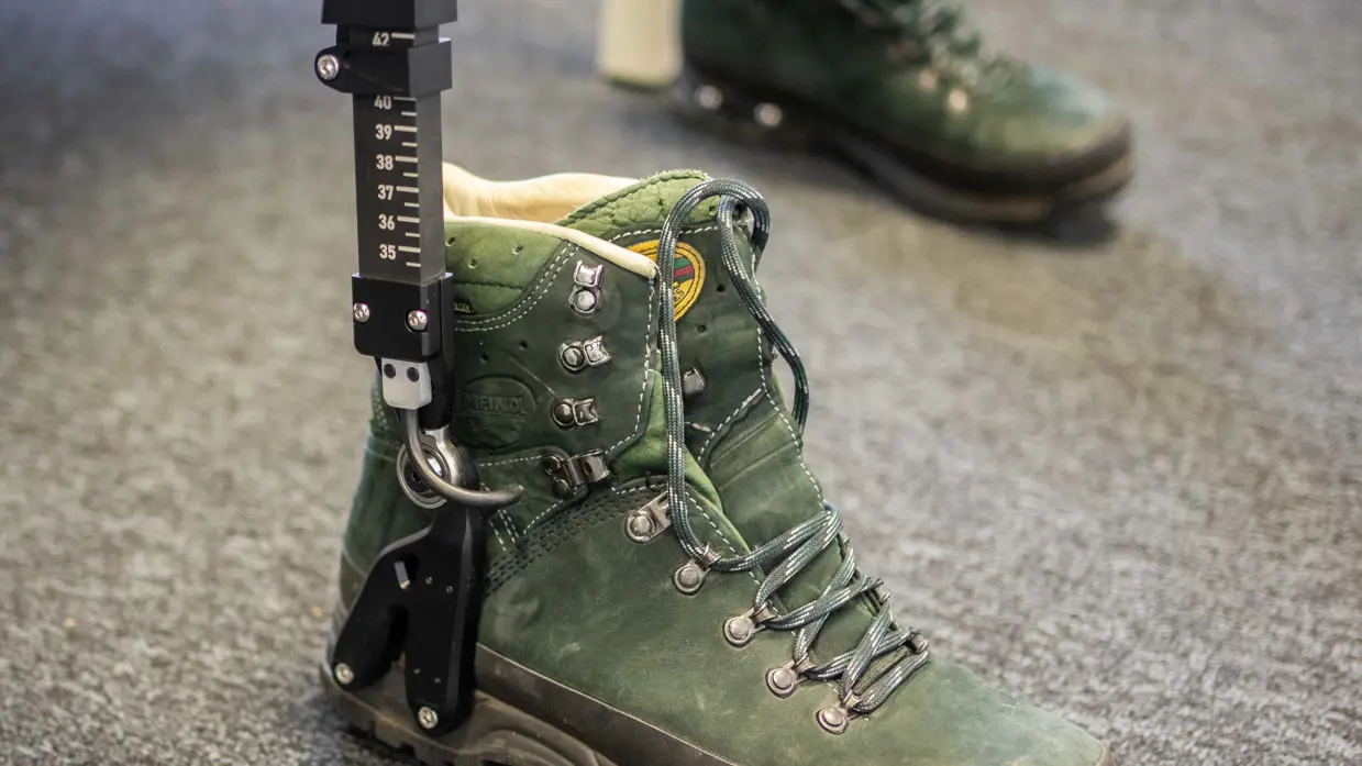 Links: een exoskelet met camouflage rugzak en groene bergschoenen staat op een tafel in een klaslokaal met stoelen en tafels.    Rechts een groene bergschoen met een zwarte metalen meetlat ernaast staat op een grijze vloer, tweede schoen op de achtergrond. 