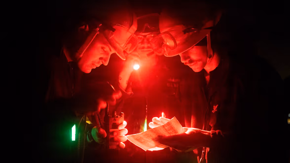 Drie personen met veiligheidsbrillen en hoofdlampen bekijken in een donkere, rood verlichte ruimte een document, terwijl een zaklamp extra licht geeft.