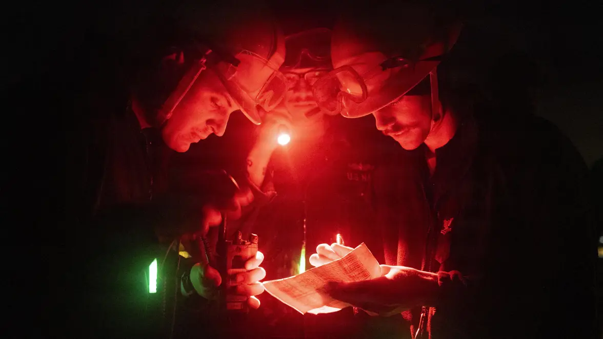 Drie personen met veiligheidsbrillen en hoofdlampen bekijken in een donkere, rood verlichte ruimte een document, terwijl een zaklamp extra licht geeft.