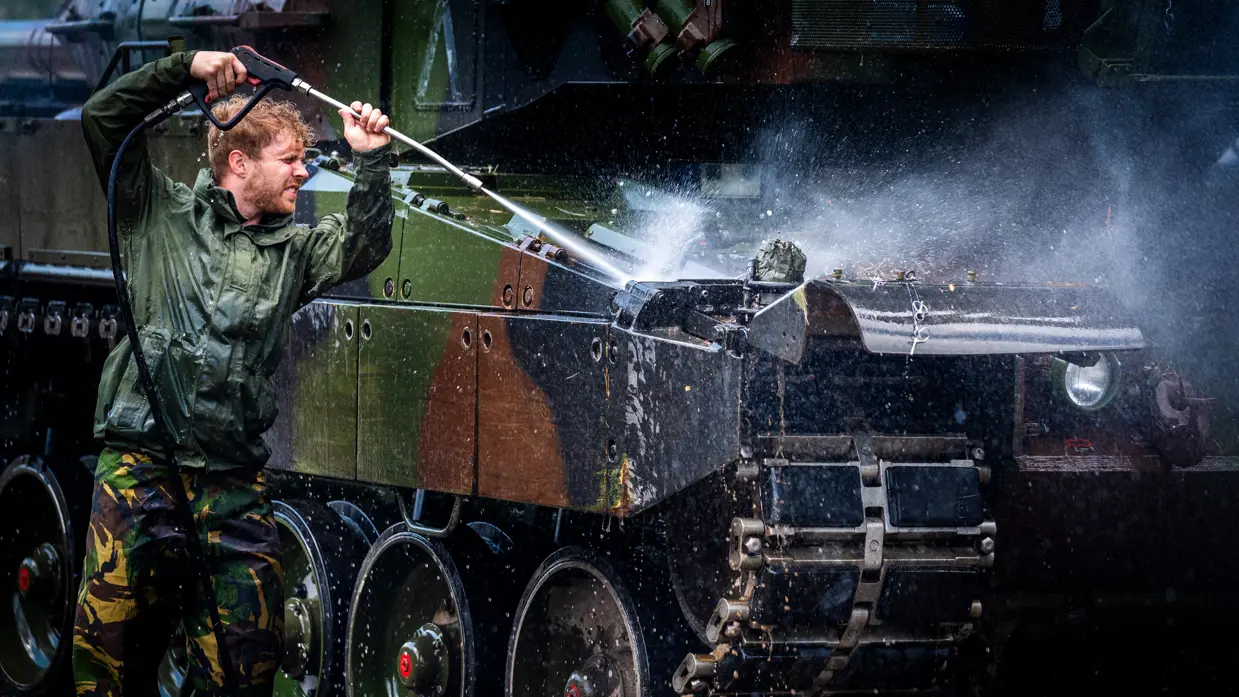 Een soldaat in een donkere jas spuit met een hogedrukreiniger water op een pantservoertuig in een bosrijke omgeving.