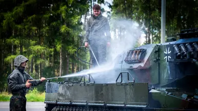 Een soldaat in een donkere jas spuit met een hogedrukreiniger water op een pantservoertuig in een bosrijke omgeving.