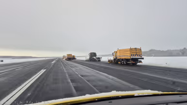 Links: drie gele sneeuwruimvoertuigen werken op een besneeuwde startbaan van een luchthaven, met een grijze lucht en sneeuw aan weerszijden. Rechts: Een Airbus 330MRTT staat op de besneeuwde luchthaven, met een oranje trap die naar de deur rijdt en sneeuwhopen op de voorgrond.