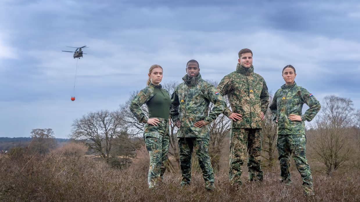 Twee vrouwelijke en twee mannelijke militairen poseren in de nieuwe kleding met het groene camouflagepatroon. Ze staan op de hei, op de achtergrond vliegt een Chinook met daaronder een oranje waterzak.