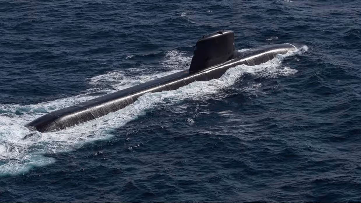 Een zwarte, stroomlijnvormige onderzeeboot vaart door golvend, donkerblauw zeewater, met witte golven achter de romp en een kleine toren bovenop. 