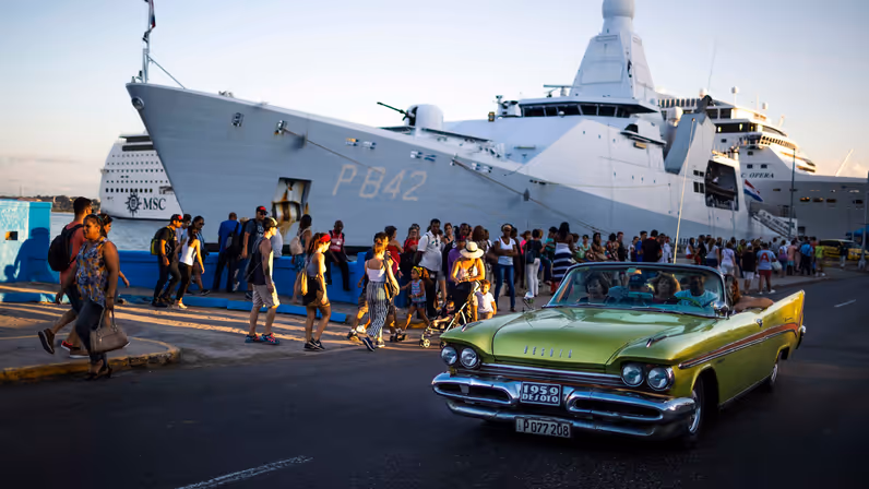 Een grijs oorlogsschip ligt tegen de kant, met matrozen voor de valreep en een blauwe antieke auto op de voorgrond.